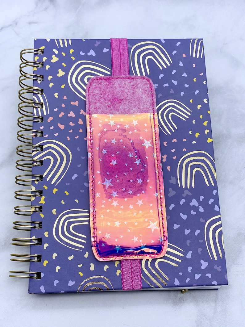 May include: A purple notebook with a gold spiral binding and a pink and iridescent star-patterned bookmark. The notebook has a rainbow and polka dot pattern.
