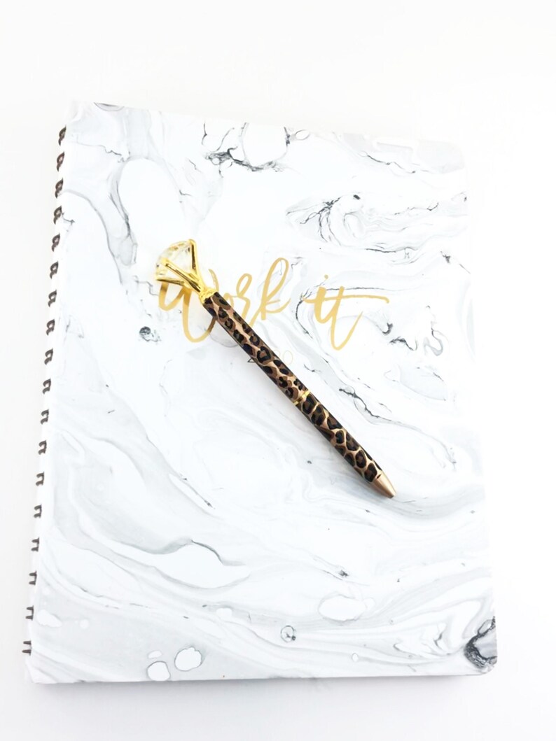 May include: A white marble notebook with a gold spiral binding and the words "Work It" written in gold script. A gold and leopard print pen is lying on top of the notebook.