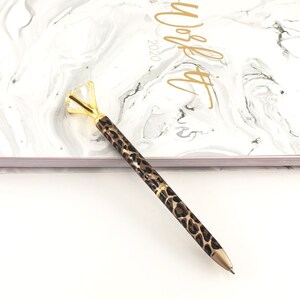 May include: A gold and black leopard print pen with a clear diamond-shaped top. The pen is lying on a white surface.