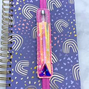 May include: A pink and iridescent pen holder with stars and a rainbow design. The holder is attached to a pink elastic band and is holding a silver pen. The holder is on top of a purple notebook with a rainbow pattern.