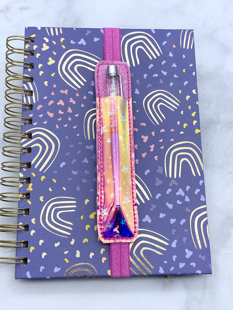 May include: A pink and iridescent pen holder with stars and a rainbow design. The holder is attached to a pink elastic band and is holding a silver pen. The holder is on top of a purple notebook with a rainbow pattern.