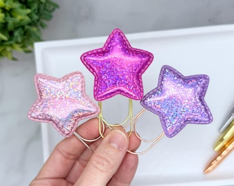 Può includere: Tre fermagli per carta a forma di stella in glitter rosa, viola e lavanda. Le clip a stella sono attaccate a fermagli per carta in metallo color oro. Le clip sono tenute in una mano.