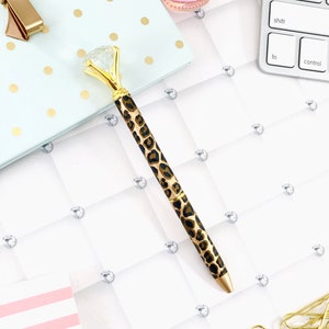 May include: A gold pen with a clear diamond-shaped top and a leopard print design. The pen is lying on a white surface with gold polka dots.