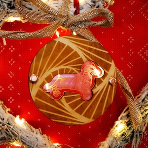 May include: A heart-shaped Christmas wreath with a wooden ornament featuring a dachshund dog. The wreath is made of frosted pine branches and fairy lights, suspended against a red patterned background.