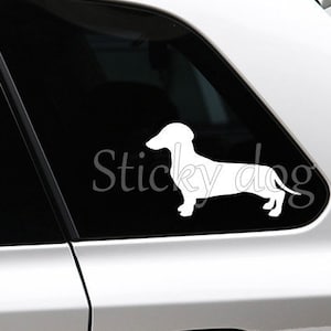 May include: A white silhouette of a dachshund dog on a silver car window. The sticker says "Sticky Dog" in white text.