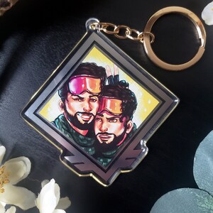 May include: A silver and gold diamond-shaped keychain with a colorful illustration of two people wearing ski goggles and winter clothing. The keychain has a gold chain and a gold ring.