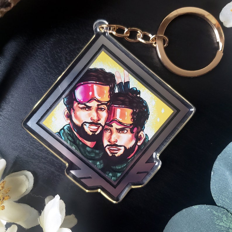 May include: A silver and gold diamond-shaped keychain with a colorful illustration of two people wearing ski goggles and winter clothing. The keychain has a gold chain and a gold ring.