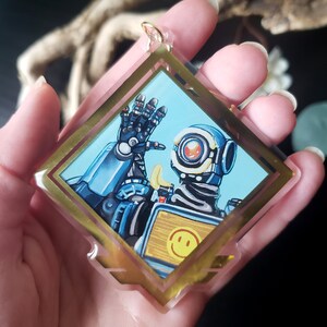 May include: A gold-framed acrylic charm featuring a cartoon illustration of a robot character from the video game Apex Legends. The robot is blue and white with a yellow smiley face on its chest. The robot is waving with its right hand.