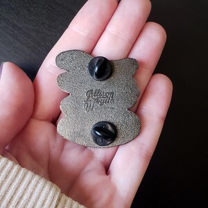 May include: Back of a silver-colored enamel pin with two black pin backs. The pin has a unique shape with rounded edges. The artist's signature, "Allison Wojda", is visible in the center of the pin.