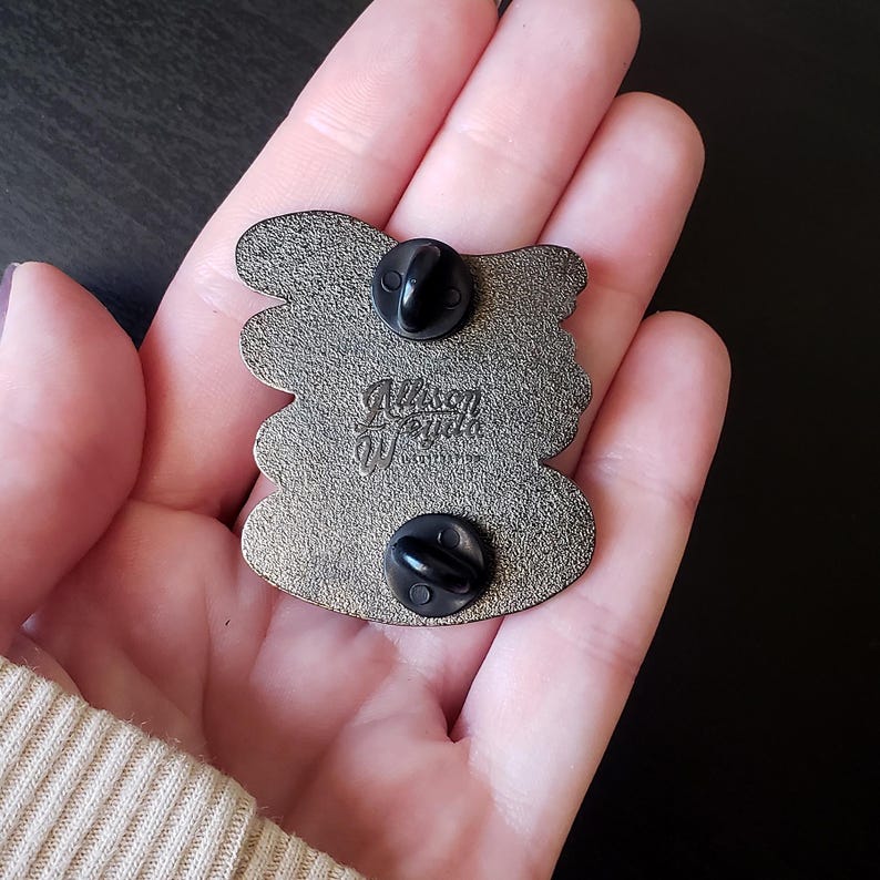 May include: Back of a silver-colored enamel pin with two black pin backs. The pin has a unique shape with rounded edges. The artist's signature, "Allison Wojda", is visible in the center of the pin.