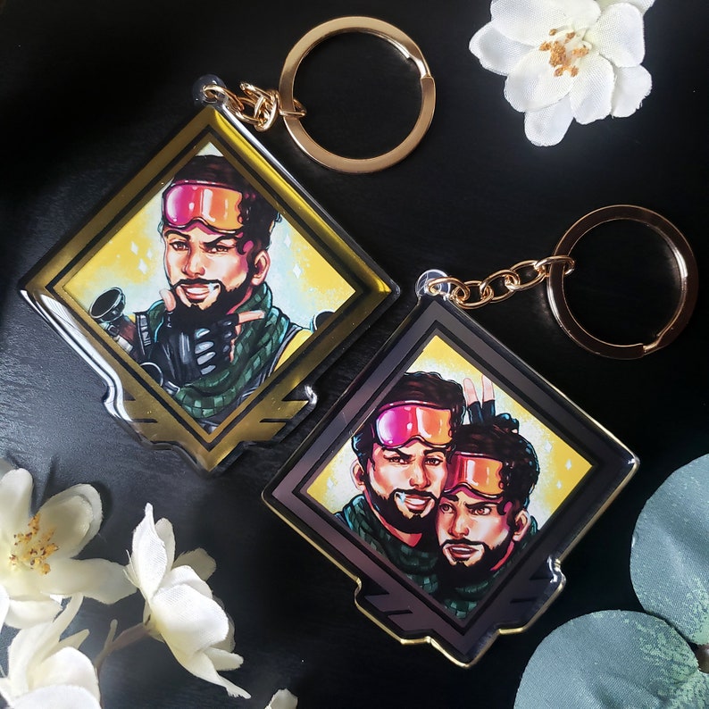 May include: Two gold keychains with diamond-shaped acrylic charms featuring illustrations of two men wearing ski goggles and winter clothing. The charms have a gold border and a black background.