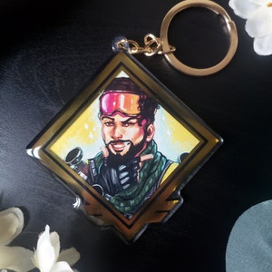 May include: A gold-colored keychain with a diamond-shaped charm featuring a cartoon illustration of a character from the video game Apex Legends. The character is wearing a pink and black ski mask, a green and brown jacket, and a black glove. The character is pointing with their right hand and has a beard and mustache.