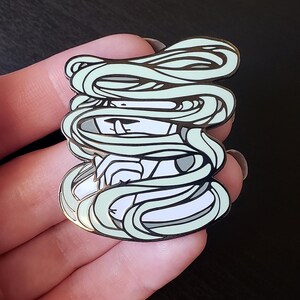 May include: A detailed enamel pin featuring a stylized mummy design. The pin has a white base with a light green wrappings design and black outlines. The mummy's face is visible, with a neutral expression. The pin is held in a hand.