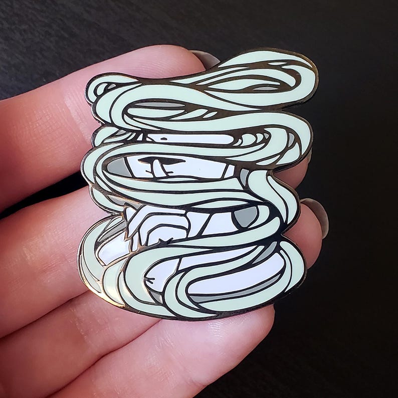 May include: A detailed enamel pin featuring a stylized mummy design. The pin has a white base with a light green wrappings design and black outlines. The mummy's face is visible, with a neutral expression. The pin is held in a hand.