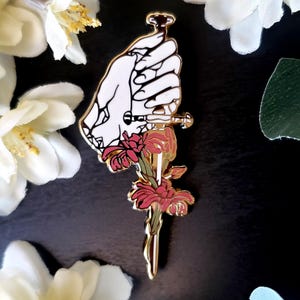 May include: A gold-colored pin featuring a white hand holding a dagger with a red flower design. The hand is detailed with black outlines, and the dagger has a floral accent. The pin is set against a dark background.