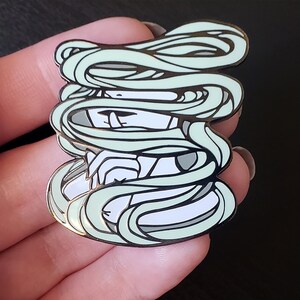 May include: Enamel pin featuring a stylized mummy design. The pin has a white and gray face with mint green wrappings. The pin is outlined in gold and has a glossy finish. The pin is a unique accessory.