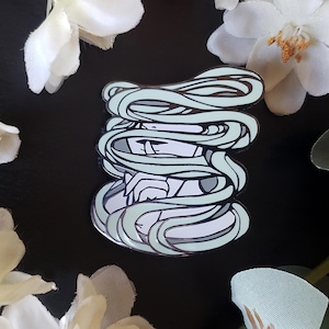 May include: A detailed enamel pin featuring a stylized figure with flowing, light teal hair. The pin has a silver outline and is set against a black background, with white floral accents.