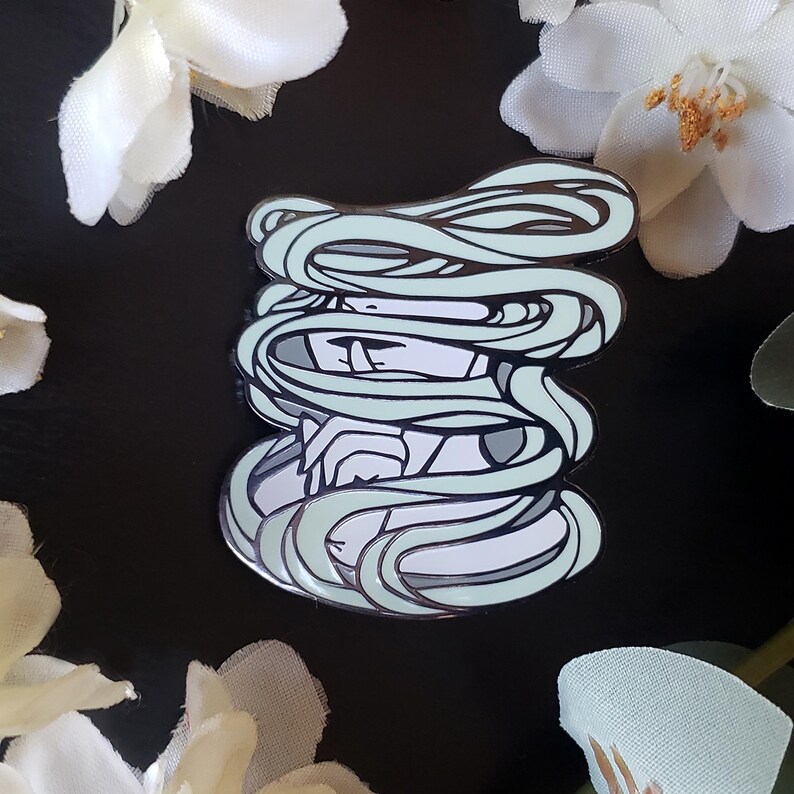 May include: A detailed enamel pin featuring a stylized figure with flowing, light teal hair. The pin has a silver outline and is set against a black background, with white floral accents.