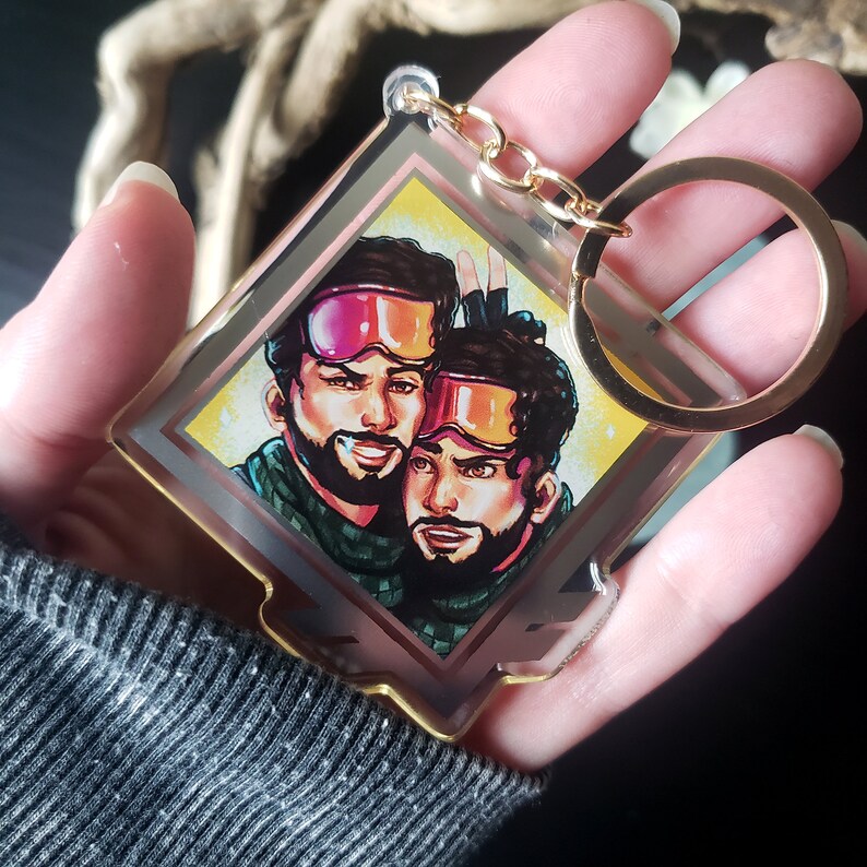 May include: A gold-colored keychain with a clear acrylic charm featuring a cartoon illustration of two people wearing ski goggles and winter clothing. The charm is shaped like a square with rounded corners and has a gold-colored border.