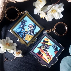 May include: Two gold and silver keychains featuring characters from the video game Apex Legends. The first keychain features a robot character with a blue and yellow color scheme. The second keychain features a human character wearing a brown fedora and a trench coat. Both keychains are diamond shaped.