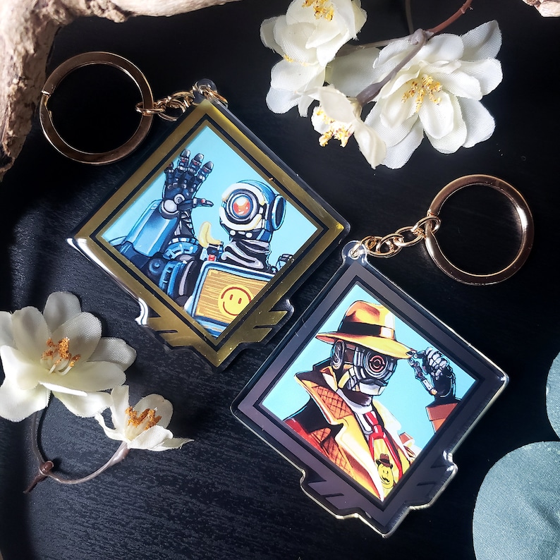 May include: Two gold and silver keychains featuring characters from the video game Apex Legends. The first keychain features a robot character with a blue and yellow color scheme. The second keychain features a human character wearing a brown fedora and a trench coat. Both keychains are diamond shaped.