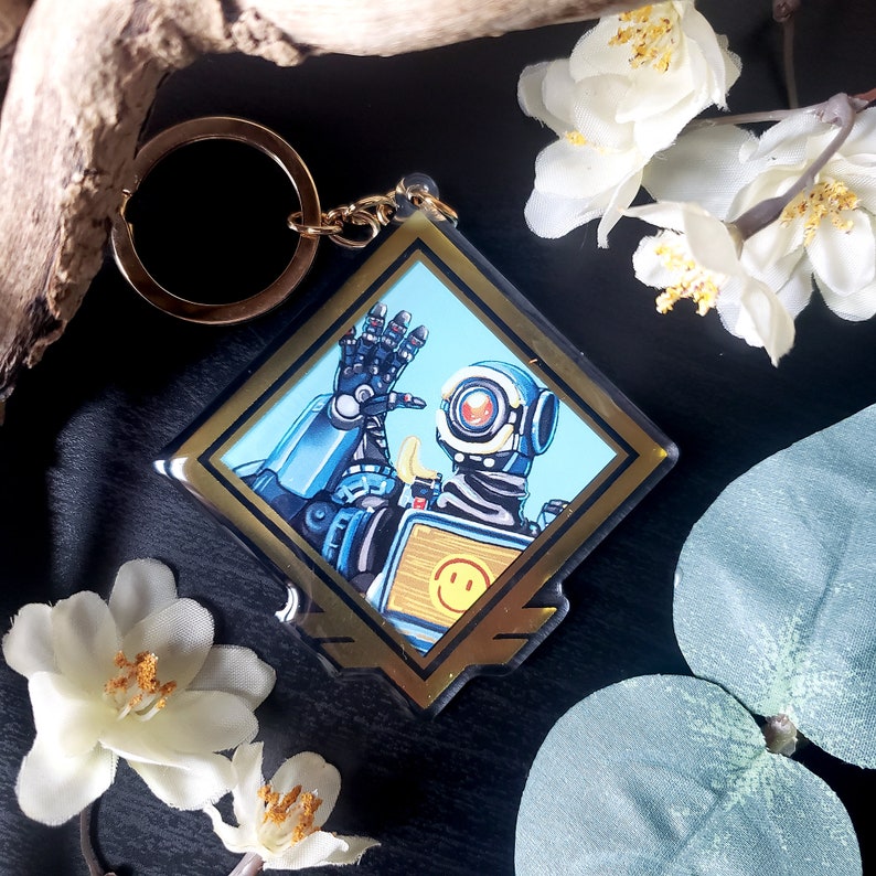 May include: A gold-framed keychain featuring a cartoon robot with a blue and white body, a yellow chest panel with a smiley face, and a single eye. The robot is holding up its right hand in a wave.