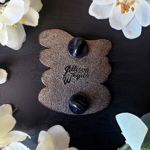 May include: A detailed close-up of the back of a pin with two black pin backs. The pin has a textured, metallic finish and a unique shape. The text "Allison Weygand Illustration" is visible on the pin.