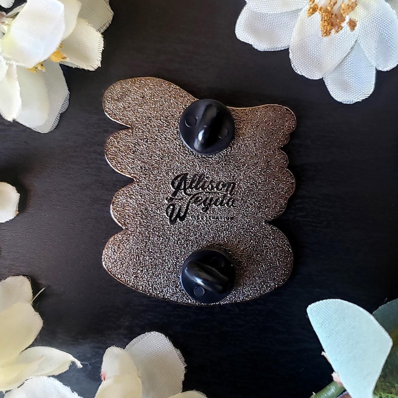 May include: A detailed close-up of the back of a pin with two black pin backs. The pin has a textured, metallic finish and a unique shape. The text "Allison Weygand Illustration" is visible on the pin.