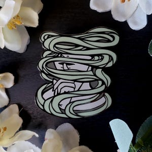 May include: A metal enamel pin featuring a stylized mummy design. The mummy is wrapped in light green bandages, with a gray face and hands visible. The pin has a black outline and is set against a dark background.