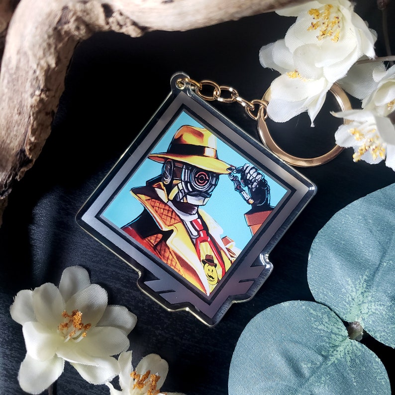May include: A silver and gold keychain featuring a cartoon character wearing a yellow fedora, a yellow trench coat, and a red tie. The character has a robotic eye and is holding a magnifying glass. The keychain is in a diamond-shaped frame.