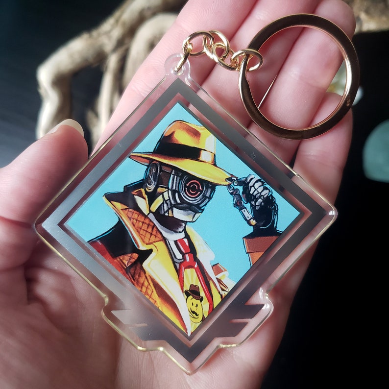May include: A clear acrylic keychain with a gold chain and a diamond-shaped design. The keychain features a cartoon illustration of a detective with a yellow fedora, a trench coat, and a robotic face. The detective is holding a magnifying glass in his right hand.