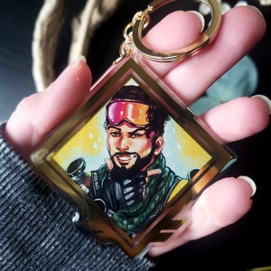 May include: A gold-colored keychain with a square-shaped charm featuring a cartoon character with a pink and black ski mask, a black beard, and a green and black jacket. The character is holding a gun and has a smirk on their face.