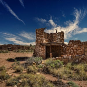 May include: A weathered stone building with a partially collapsed roof stands in a desert landscape. The sky is a bright blue with wispy white clouds.
