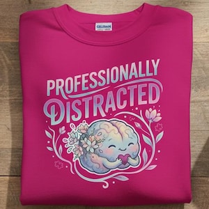 Può includere: Una felpa magenta con la scritta "Professionally Distracted" e una grafica di un cervello dei cartoni animati. Il cervello è circondato da fiori e tiene un cuore. La felpa è su una superficie di legno con un taccuino, una penna, occhiali e una tazza di caffè.