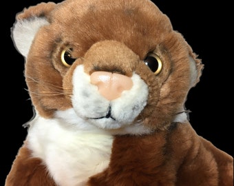 May include: A soft toy cougar with brown fur, a white chest, and a pink nose. The cougar has large, expressive eyes and a detailed face, giving it a realistic appearance. The toy is designed to resemble a wild cat.