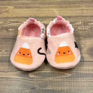 Puede incluir: Un par de botines de bebé rosas con un diseño de caramelo de maíz. Los botines tienen un patrón de gato negro y fantasma en los lados.