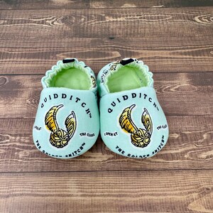 May include: A pair of baby booties in a light teal color, featuring a gold and black graphic of a winged ball. The text "QUIDDITCH" and "THE GOLDEN SNITCH" are printed on the booties.