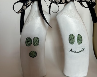 Puede incluir: Dos botellas blancas con forma de fantasma con ojos y bocas verdes, decoradas con una cinta negra con un patrón de fantasma blanco.