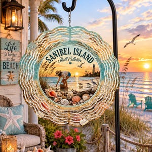 May include: A circular metal wind spinner featuring a beach scene with a person collecting shells, a lighthouse, and the text Sanibel Island Shell Collector. The design includes a scalloped edge with a gold and teal finish, hanging from a black hook.
