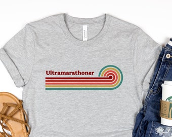 May include: A gray t-shirt with the text "Ultramarathoner" in red and a retro rainbow design. The shirt is laid flat on a white surface with a pair of brown sandals and blue jeans with a coffee cup in the pocket.
