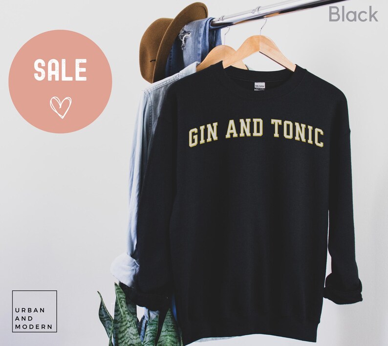 May include: Black sweatshirt with the text "GIN AND TONIC" in a gold, collegiate-style font. The sweatshirt is hanging on a wooden hanger with other clothing items. A "SALE" sign is in the upper left corner.