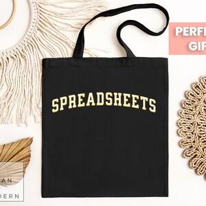 May include: Black tote bag with the word "SPREADSHEETS" in gold arched lettering. The bag has black handles and is made of a sturdy fabric. The words "URBAN AND MODERN" are in a white box in the lower left corner.