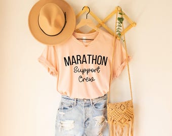 May include: A peach-colored t-shirt with the words "MARATHON Support Crew" in black script. The shirt is paired with distressed denim shorts, a tan hat, and a woven crossbody bag. The items are displayed on a wooden hanger against a cream-colored wall.