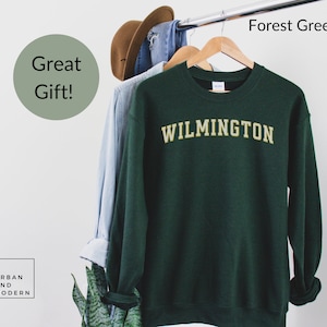 May include: Forest green sweatshirt with the word "WILMINGTON" in gold lettering. The sweatshirt is hanging on a wooden hanger with a light blue shirt and a brown hat. The words "Great Gift!" are in a green circle.