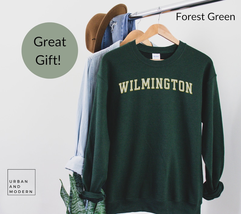 May include: Forest green sweatshirt with the word "WILMINGTON" in gold lettering. The sweatshirt is hanging on a wooden hanger with a light blue shirt and a brown hat. The words "Great Gift!" are in a green circle.