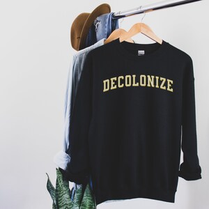 Pu&ograve; includere: Felpa nera con la scritta "DECOLONIZE" in lettere dorate.