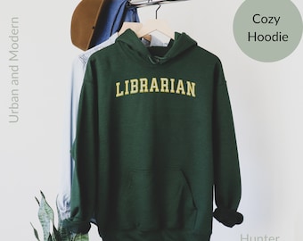 May include: A hunter green hoodie with the word "LIBRARIAN" in yellow lettering. The hoodie is hanging on a rack with a hat and a shirt.
