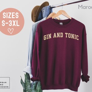May include: A maroon sweatshirt with the text "GIN AND TONIC" in a gold, collegiate-style font. The sweatshirt is hanging on a wooden hanger. The sizes available are S-3XL.