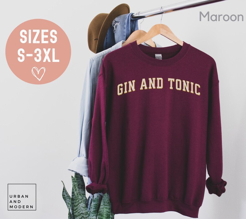 May include: A maroon sweatshirt with the text "GIN AND TONIC" in a gold, collegiate-style font. The sweatshirt is hanging on a wooden hanger. The sizes available are S-3XL.
