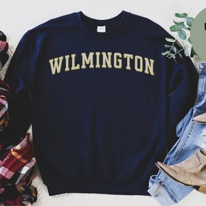 May include: Navy blue sweatshirt with "WILMINGTON" in gold lettering. The image includes a plaid scarf, jeans, and boots. The sweatshirt is labeled "Unisex sizing". The brand name "URBAN AND MODERN" is also visible.